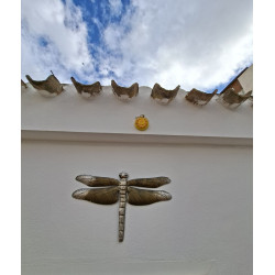 Adorno de flor de cemento amarilla colgado en fachada blanca bajo tejas rústicas junto a gran libélula decorativa.