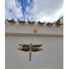 Adorno de flor de cemento amarilla colgado en fachada blanca bajo tejas rústicas junto a gran libélula decorativa.