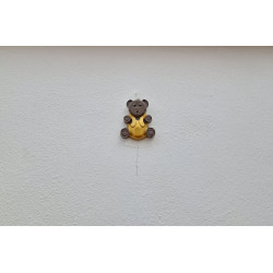 Adorno de osito grande de cemento con peto amarillo pintado a mano colgado de forma vertical en una pared blanca de exterior.