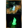 Adorno Papá Noel de cemento fosforescente con gorro inclinado de día en el abeto y brillando verde de noche.