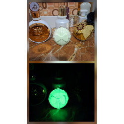 Adorno bola Navidad de cemento fosforescente con diseño de regalo y lazo de día junto a perfumes y brillo verde en la oscuridad.