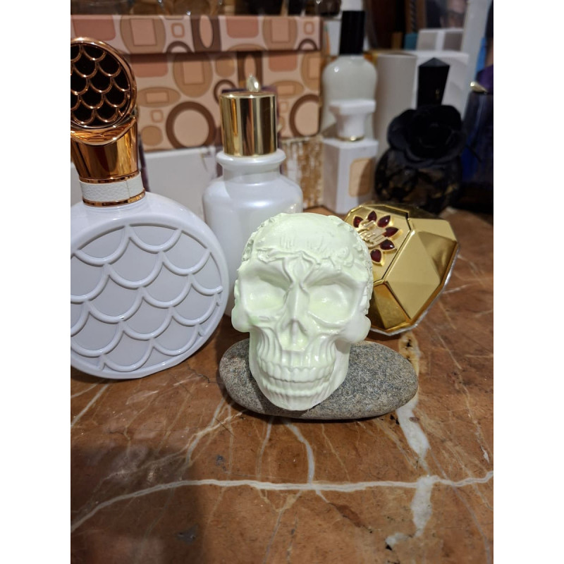 Adorno de calavera de cemento fosforescente con corona de espinas de día apoyada en una piedra junto a perfumes sobre mármol.